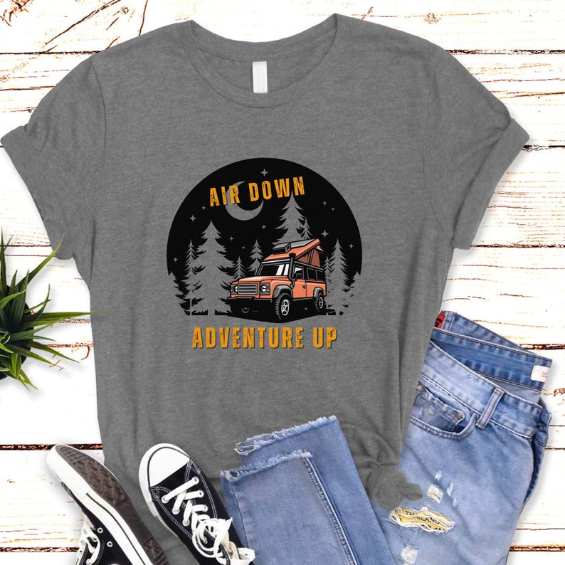 Puede incluir: Camiseta gris con un gr&aacute;fico de un coche en un bosque de noche. El texto "Air Down Adventure Up" est&aacute; impreso en la camiseta.