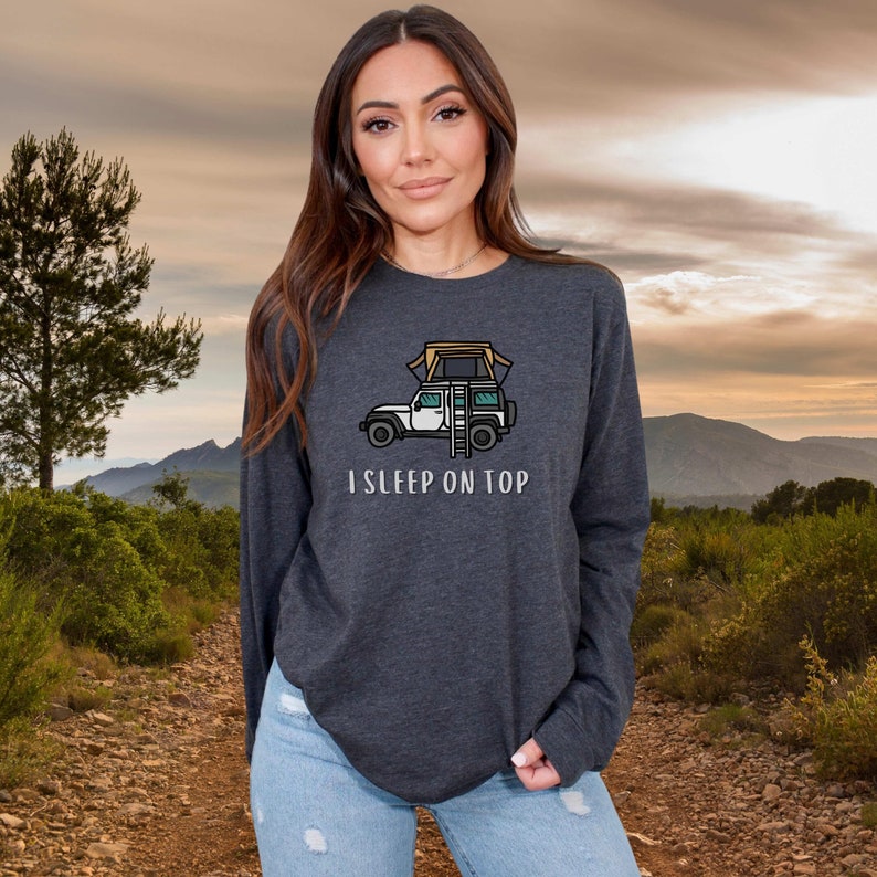 May include: A dark heather grey long-sleeved shirt with a cartoon illustration of a car with a rooftop tent and the text "I SLEEP ON TOP".