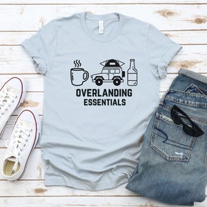 Puede incluir: Una camiseta azul claro con un gr&aacute;fico negro de una taza de caf&eacute;, un coche con una tienda de campa&ntilde;a en la parte superior y una botella de cerveza. El texto "Overlanding Essentials" est&aacute; impreso debajo del gr&aacute;fico.