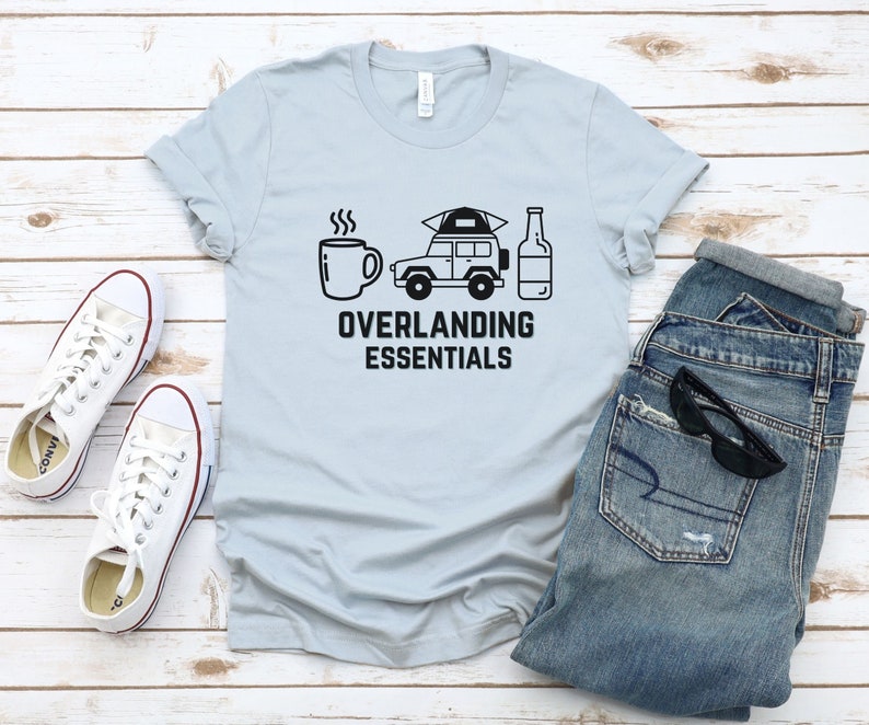 Puede incluir: Una camiseta azul claro con un gr&aacute;fico negro de una taza de caf&eacute;, un coche con una tienda de campa&ntilde;a en la parte superior y una botella de cerveza. El texto "Overlanding Essentials" est&aacute; impreso debajo del gr&aacute;fico.
