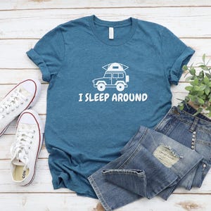 Pu&ograve; includere: Una maglietta blu turchese con una grafica bianca di un'auto con una tenda sul tetto. Il testo "I SLEEP AROUND" &egrave; stampato sotto l'auto.