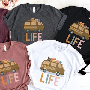 May include: A collection of t-shirts in various colors, each featuring a brown van graphic with luggage and the word "LIFE" in colorful letters. The shirts are made of a soft material.
