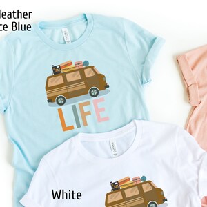 May include: Light blue t-shirt with a brown camper van graphic and the word "LIFE" in orange, gray, and pink. The van has luggage on top. The shirt is made of a soft material.