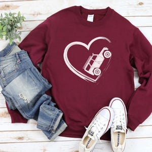 May include: Burgundy sweatshirt with a white heart design featuring a vehicle. The sweatshirt is paired with blue jeans and white sneakers. The design is a simple line drawing of a vehicle inside a heart.