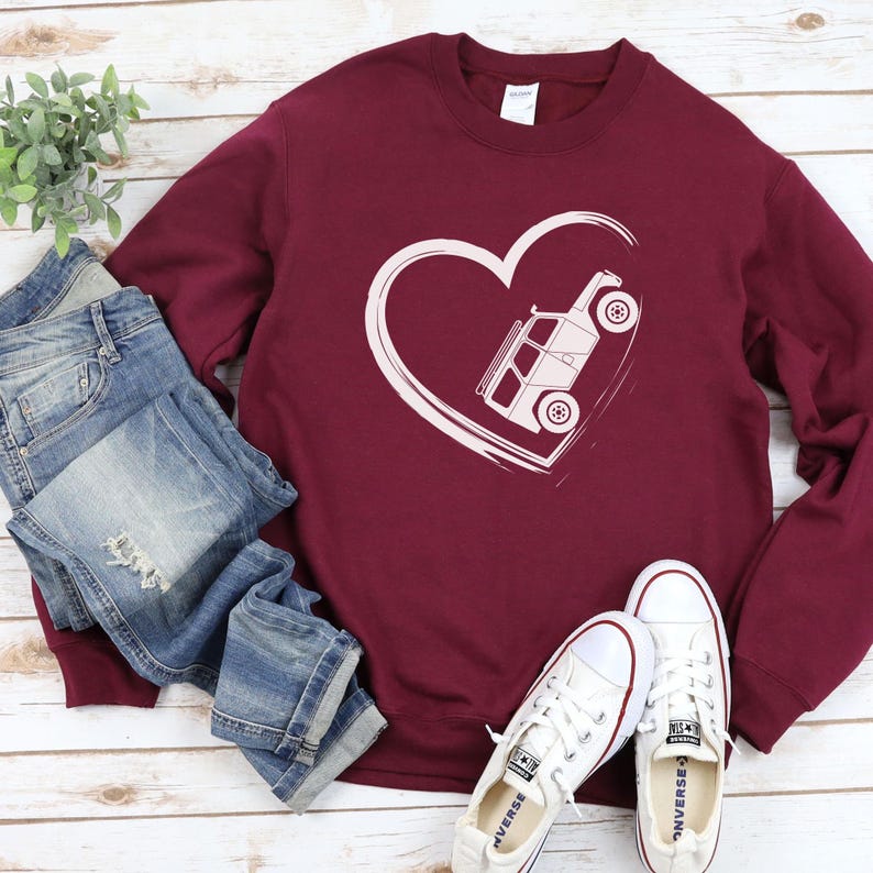 May include: Burgundy sweatshirt with a white heart design featuring a vehicle. The sweatshirt is paired with blue jeans and white sneakers. The design is a simple line drawing of a vehicle inside a heart.
