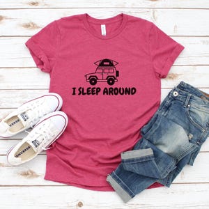 Pu&ograve; includere: Una maglietta rosa con una grafica nera di un'auto con una tenda sul tetto e il testo "I sleep around". La maglietta &egrave; appoggiata su una superficie di legno con un paio di scarpe da ginnastica bianche e un paio di jeans blu.
