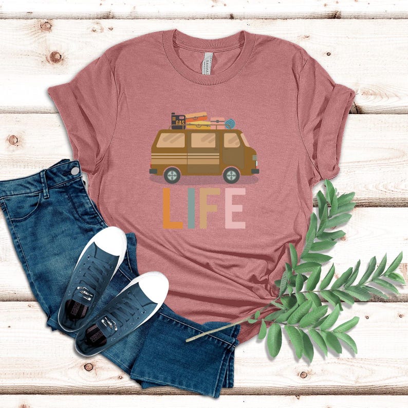 May include: A dusty rose t-shirt featuring a brown camper van graphic with luggage on top and the word "LIFE" in colorful letters. The shirt is paired with blue jeans and navy sneakers. A sprig of greenery is on the right.