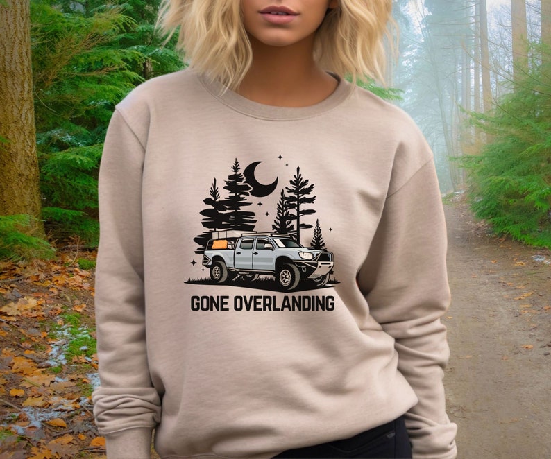 May include: A tan sweatshirt with a graphic of a truck with a camper shell driving through a forest at night. The text "Gone Overlanding" is printed below the truck.