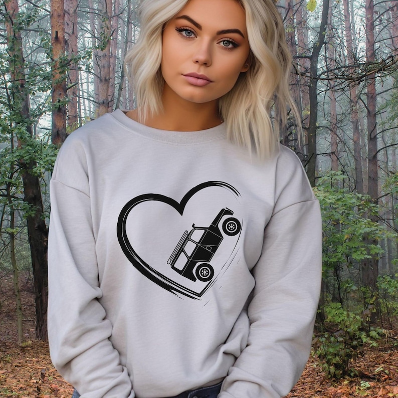 May include: A light gray sweatshirt featuring a black heart with a black off-road vehicle graphic inside. The sweatshirt has long sleeves and a crew neck. The background is a forest scene.