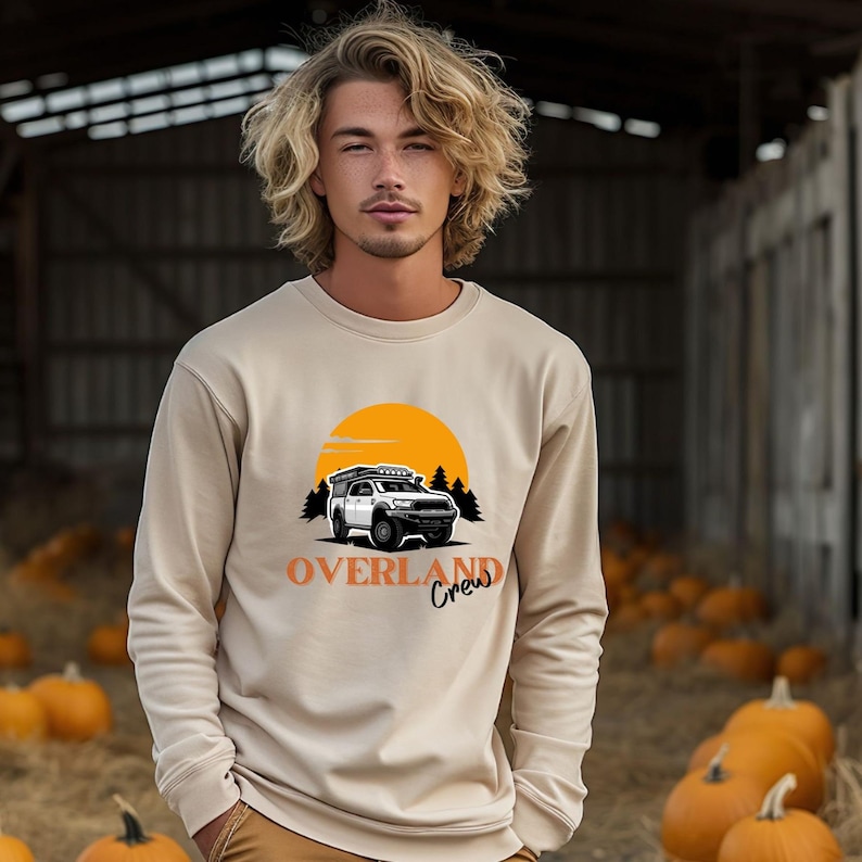 May include: A beige sweatshirt featuring a graphic of a white truck with a rooftop tent driving through a forest with a yellow sun in the background. The text "Overland Crew" is printed below the truck.