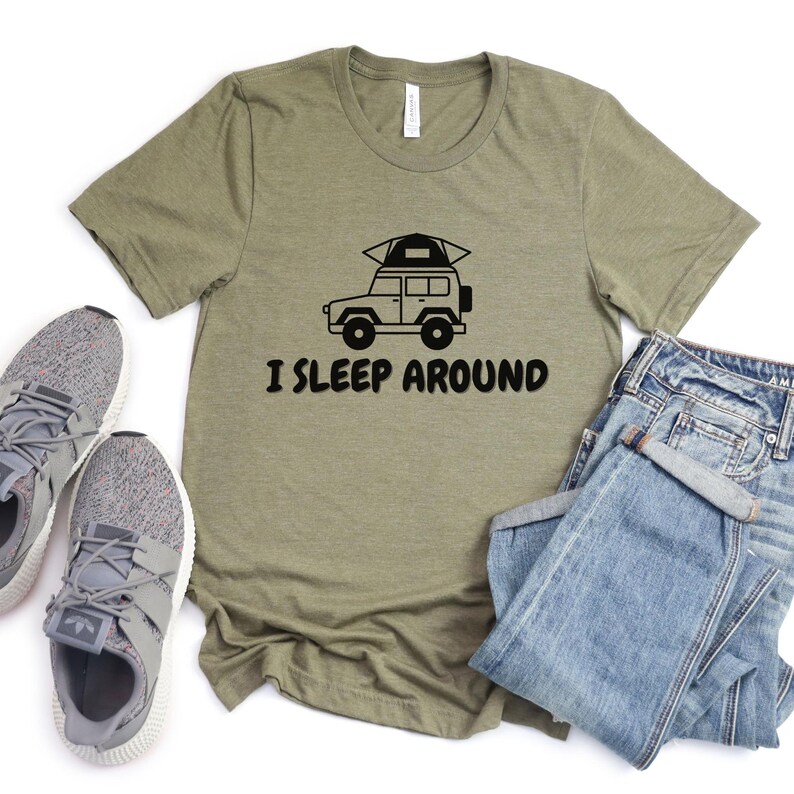 Pu&ograve; includere: Una maglietta verde con una grafica nera di un'auto con una tenda sul tetto. Il testo "I SLEEP AROUND" &egrave; stampato sotto l'auto.
