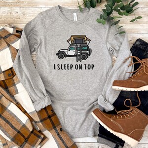 May include: A gray long-sleeved shirt with a white Jeep with a tent on top and the text "I SLEEP ON TOP".
