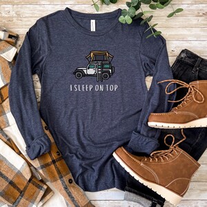 May include: A navy blue long-sleeved t-shirt with a white jeep and a tent on top. The text "I SLEEP ON TOP" is printed below the jeep.