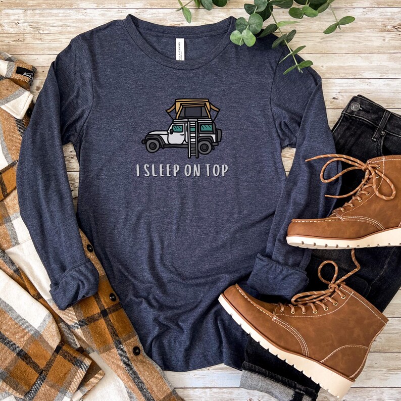 May include: A navy blue long-sleeved t-shirt with a white jeep and a tent on top. The text "I SLEEP ON TOP" is printed below the jeep.
