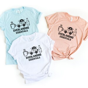 Puede incluir: Tres camisetas con un gr&aacute;fico en blanco y negro de un coche con una tienda de campa&ntilde;a en el techo, una taza de caf&eacute; y una botella de cerveza. El texto en las camisetas dice "Overlanding Essentials". Las camisetas son de color azul claro, melocot&oacute;n y blanco.