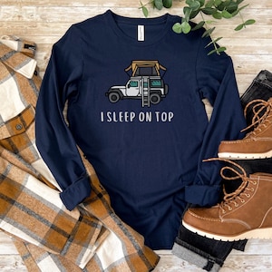 May include: A navy blue long-sleeved t-shirt with a white graphic of a car with a rooftop tent and the text "I SLEEP ON TOP".