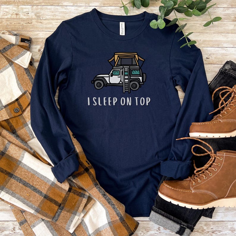 May include: A navy blue long-sleeved t-shirt with a white graphic of a car with a rooftop tent and the text "I SLEEP ON TOP".