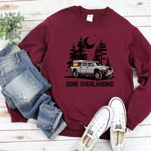 May include: A maroon sweatshirt with a graphic of a truck with a rooftop tent driving through a forest at night. The text "Gone Overlanding" is printed below the truck.