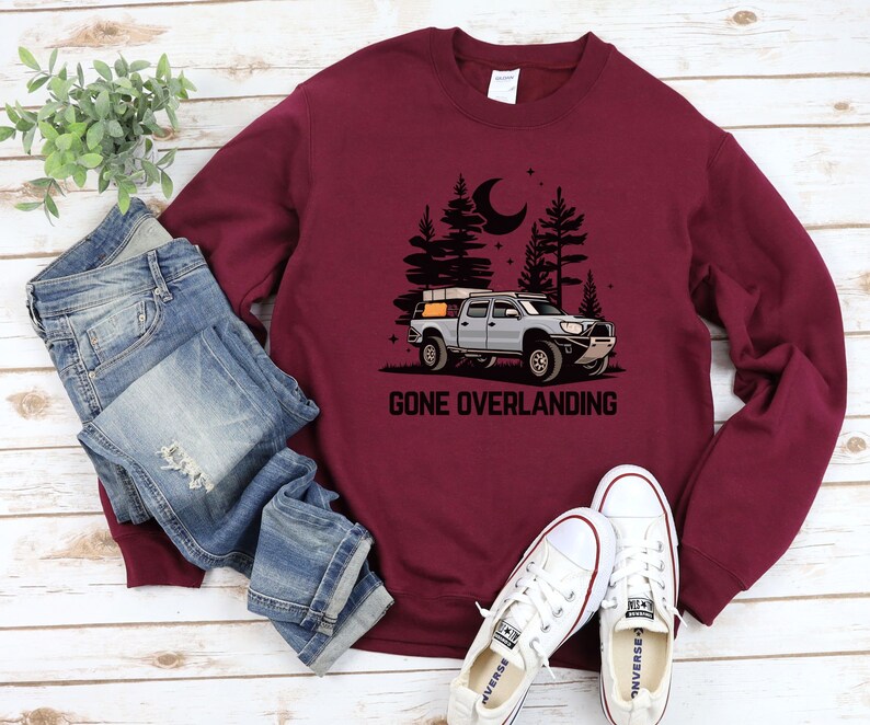 May include: A maroon sweatshirt with a graphic of a truck with a rooftop tent driving through a forest at night. The text "Gone Overlanding" is printed below the truck.