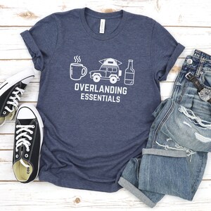 Puede incluir: Una camiseta azul marino con un gr&aacute;fico blanco de una taza de caf&eacute;, un jeep con una tienda de campa&ntilde;a en la parte superior y una botella de cerveza. El texto "Overlanding Essentials" est&aacute; impreso debajo del gr&aacute;fico.