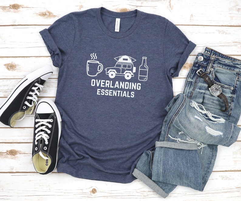 Puede incluir: Una camiseta azul marino con un gr&aacute;fico blanco de una taza de caf&eacute;, un jeep con una tienda de campa&ntilde;a en la parte superior y una botella de cerveza. El texto "Overlanding Essentials" est&aacute; impreso debajo del gr&aacute;fico.