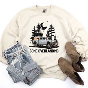 May include: A beige sweatshirt with a black and white illustration of a truck with a rooftop tent driving through a forest at night. The text "Gone Overlanding" is printed below the truck.
