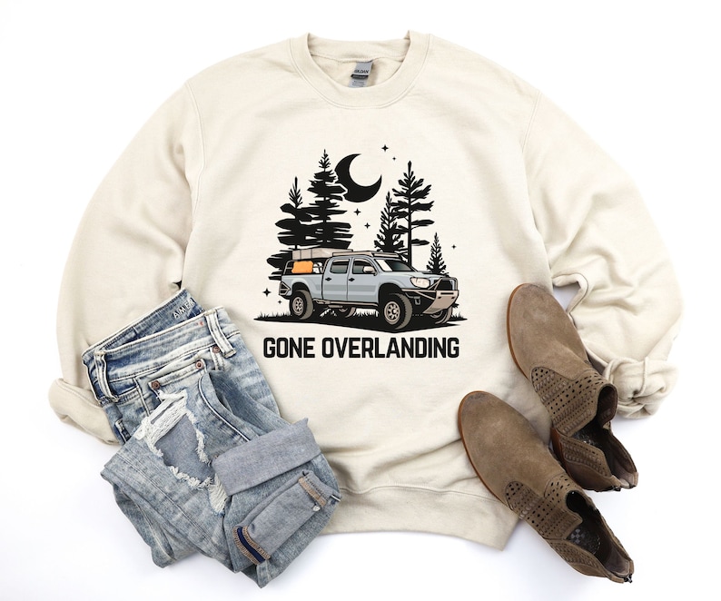 May include: A beige sweatshirt with a black and white illustration of a truck with a rooftop tent driving through a forest at night. The text "Gone Overlanding" is printed below the truck.