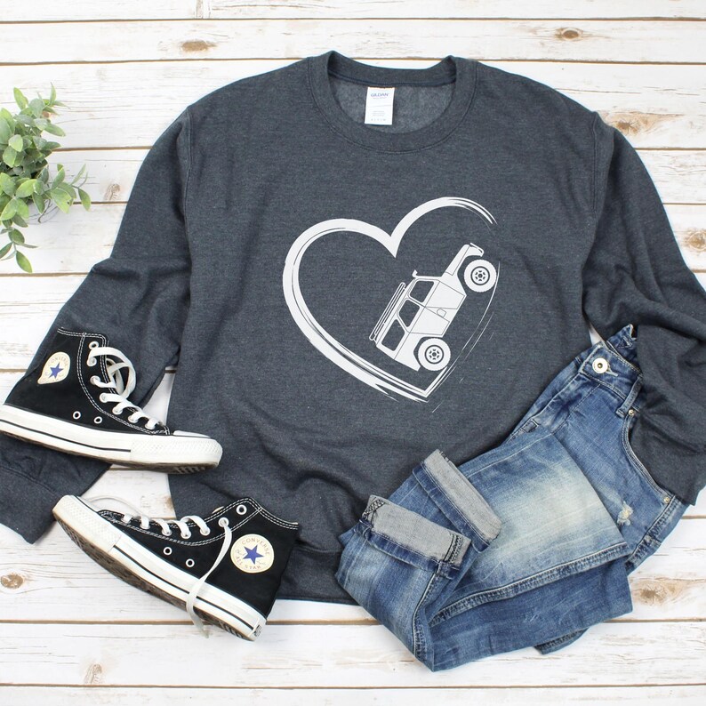 May include: A dark gray sweatshirt with a white heart design containing a white off-road vehicle. The image also shows a pair of black high-top sneakers and blue jeans. The sneakers have white laces and soles.