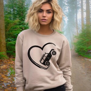 May include: A beige sweatshirt featuring a black heart-shaped design with a stylized off-road vehicle inside. The sweatshirt has long sleeves and a crew neck. The background shows a forest with a path.