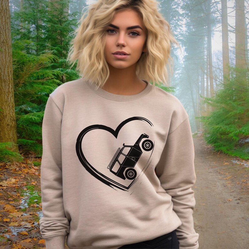 May include: A beige sweatshirt featuring a black heart-shaped design with a stylized off-road vehicle inside. The sweatshirt has long sleeves and a crew neck. The background shows a forest with a path.