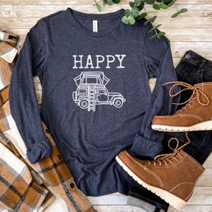 May include: A navy blue long-sleeved shirt with the word "HAPPY" printed in white on the front. A white outline of a car with a rooftop tent is below the word "HAPPY". The shirt is laid out on a wooden surface with a plaid shirt, jeans, and brown boots.