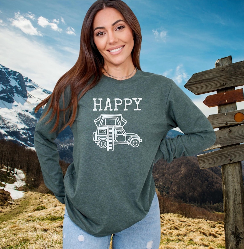 May include: A dark green long-sleeved shirt with the word "HAPPY" and a white outline of a car with a rooftop tent and ladder. The shirt is being worn by a person in front of a mountain background.