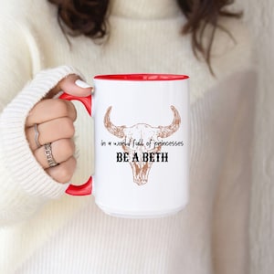 May include: White ceramic mug with a red rim and handle. The mug features a brown line drawing of a bull skull with the text "in a world full of princesses BE A BETH".