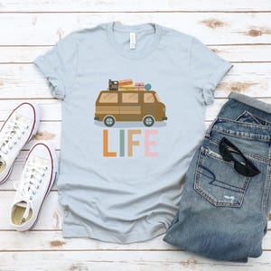 May include: Light blue t-shirt with a brown van graphic carrying luggage on top. The word "LIFE" is printed in orange, green, and pink. White sneakers and blue jeans are also in the image.