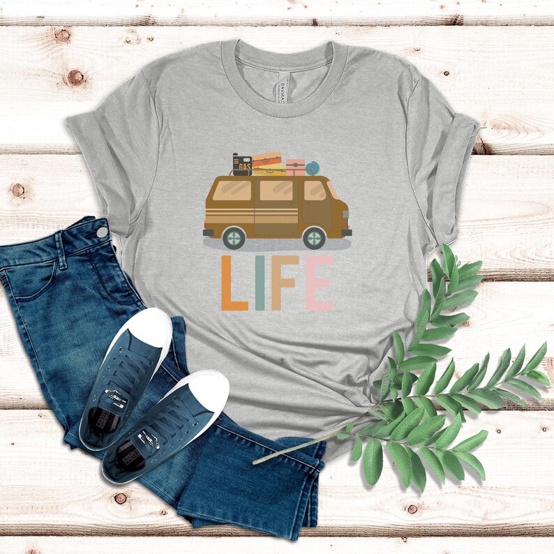 May include: A gray t-shirt with a brown van graphic carrying luggage and the word "LIFE" in colorful letters. The shirt is paired with blue jeans and sneakers. A sprig of greenery is on the right.