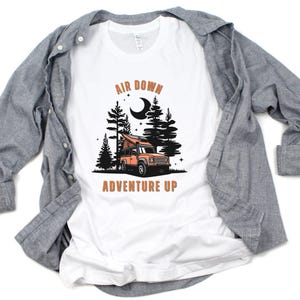 May include: White t-shirt featuring a graphic of a camper van in a forest at night. The text "Air Down Adventure Up" is printed on the shirt.
