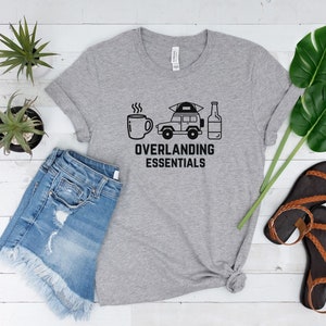 Puede incluir: Una camiseta gris con un gr&aacute;fico negro de una taza de caf&eacute;, un coche con una tienda de campa&ntilde;a en la parte superior y una botella de cerveza. El texto "Overlanding Essentials" est&aacute; debajo del gr&aacute;fico.