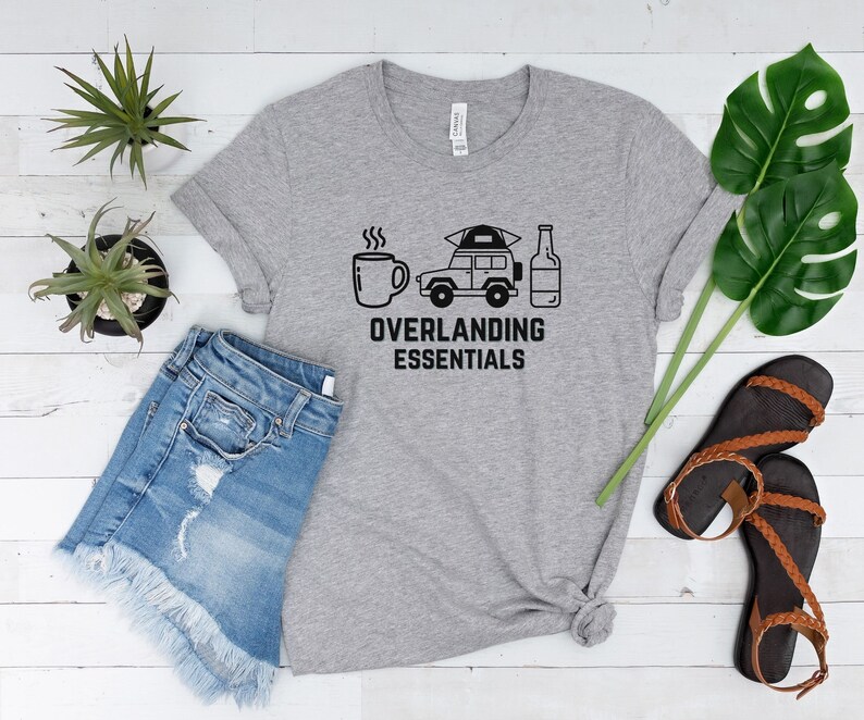 Puede incluir: Una camiseta gris con un gr&aacute;fico negro de una taza de caf&eacute;, un coche con una tienda de campa&ntilde;a en la parte superior y una botella de cerveza. El texto "Overlanding Essentials" est&aacute; debajo del gr&aacute;fico.