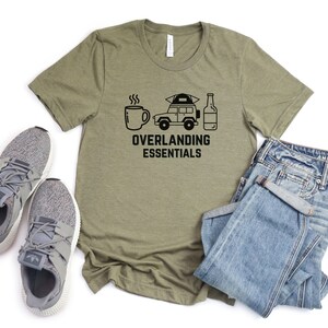 Puede incluir: Una camiseta verde con un gr&aacute;fico negro de una taza de caf&eacute;, un coche con una tienda de campa&ntilde;a en la parte superior y una botella de cerveza. El texto "Overlanding Essentials" est&aacute; debajo del gr&aacute;fico.