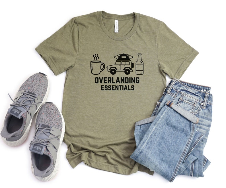 Puede incluir: Una camiseta verde con un gr&aacute;fico negro de una taza de caf&eacute;, un coche con una tienda de campa&ntilde;a en la parte superior y una botella de cerveza. El texto "Overlanding Essentials" est&aacute; debajo del gr&aacute;fico.