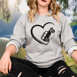 May include: A gray sweatshirt featuring a black heart with a stylized off-road vehicle inside. The vehicle is depicted at an angle, giving a sense of motion. The sweatshirt has long sleeves and a crew neck.