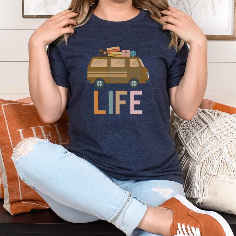 May include: Navy blue t-shirt featuring a brown camper van graphic with luggage on top. The word "LIFE" is printed below the van in colorful block letters. The shirt is made of a soft material.