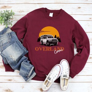 May include: A maroon sweatshirt featuring a graphic of a white truck with a roof rack driving through a sunset landscape. The text "Overland Crew" is printed below the truck.