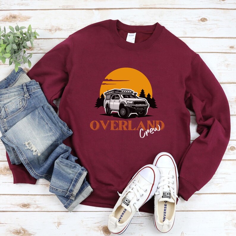 May include: A maroon sweatshirt featuring a graphic of a white truck with a roof rack driving through a sunset landscape. The text "Overland Crew" is printed below the truck.