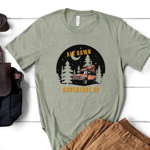 Puede incluir: Una camiseta verde con un gr&aacute;fico de un coche en un bosque de noche. El texto "Air Down Adventure Up" est&aacute; impreso en la camiseta.