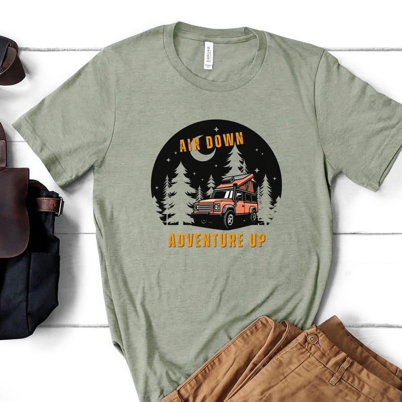 Puede incluir: Una camiseta verde con un gr&aacute;fico de un coche en un bosque de noche. El texto "Air Down Adventure Up" est&aacute; impreso en la camiseta.