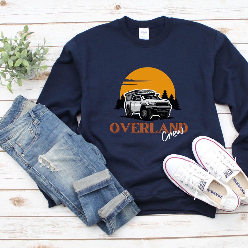 May include: A navy blue sweatshirt featuring a white truck graphic and the text "Overland Crew" in orange. The truck is driving through a forest with a sunset in the background.