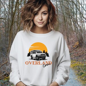 May include: White sweatshirt featuring an orange sunset graphic and a white truck with a roof rack. The text "OVERLAND Crew" is printed in orange below the truck.