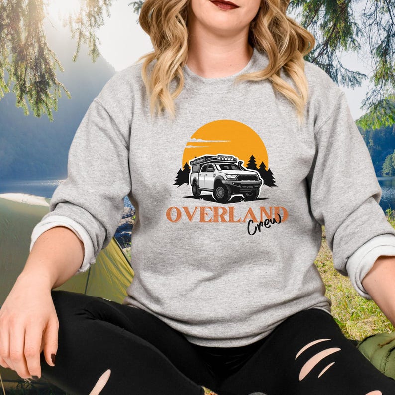 May include: A grey sweatshirt with an orange graphic of a white truck with a roof top tent driving through a forest with a yellow sun in the background. The text "Overland Crew" is printed below the truck.