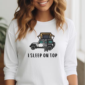 May include: White long-sleeve t-shirt with a cartoon illustration of a car with a rooftop tent and a ladder. The text "I SLEEP ON TOP" is printed below the image.